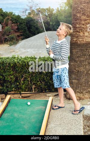 Boy enthusiastically plays mini golf on summer evening on the territory of the hotel Stock Photo
