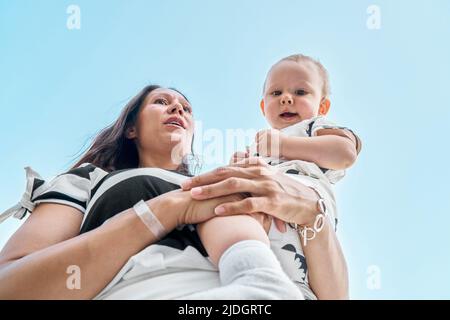 Mother holds cute smiling toddler in arms against blue sky. Young brunette woman and little boy enjoy summer vacation low angle close view Stock Photo