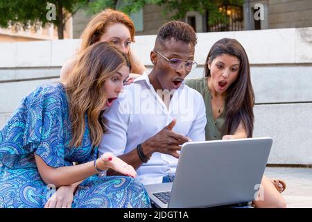 Three women and a man looking at a laptop with a surprised face in a urban park Stock Photo