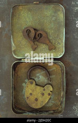 Old brass padlock lying in brass tin with its rusty keys lying in lid and resting on rusty metal sheet Stock Photo