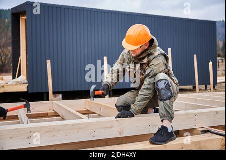 Man worker building wooden frame house on pile foundation. Carpenter hammering nail into wooden joist, using hammer. Carpentry concept. Stock Photo