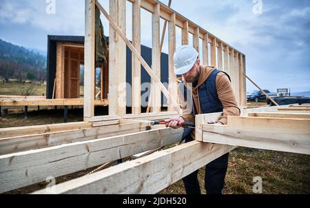 Man worker building wooden frame house on pile foundation. Carpenter hammering nail into wooden plank, using hammer. Carpentry concept. Stock Photo