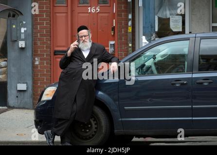 A hasidic Jewish man likely a rabbi, speaks on his cell phone while leaning up against a car. On Division Avenue in Williamsburg, Brooklyn, New York. Stock Photo