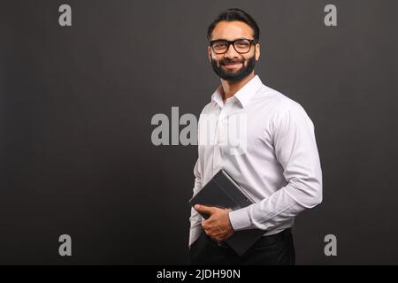 Indian male entrepreneur in formal white shirt and glasses carrying laptop isolated on black, bearded businessman using laptop, using new computer app, looking at the camera and smiles Stock Photo