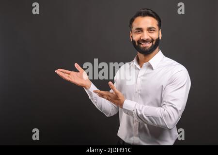 Indian male entrepreneur points with both hands aside isolated on black, bearded businessman presenting, showing novelty looking at the camera and smiles, copy space Stock Photo