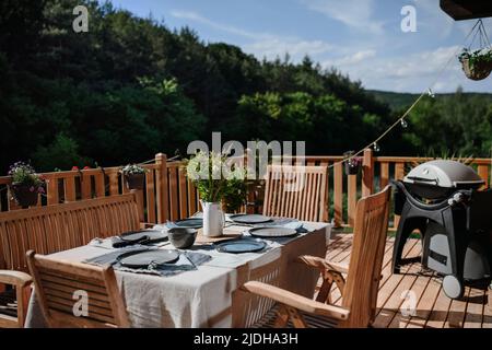 Dining table with wooden chairs set for dinner on the terrace with grill in summer, garden party. concept. Stock Photo