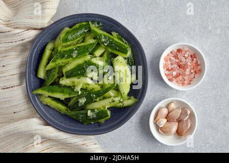 Smashed lightly salted cucumbers in a ceramic bowl surrounded by garlic, pink salt and kitchen towel napkin. Top view with copy space Stock Photo