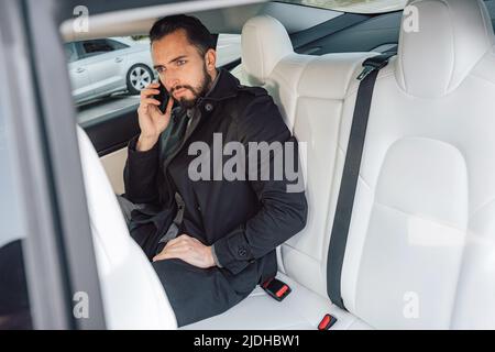 Caucasian young successful man seriously talking on the smartphone while sitting in the back seat of a driving car Stock Photo