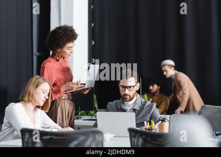 African american woman pointing at laptop near multiethnic colleagues working in ad agency Stock Photo