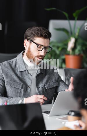 bearded designer in eyeglasses working near laptop in ad agency near blurred colleague Stock Photo