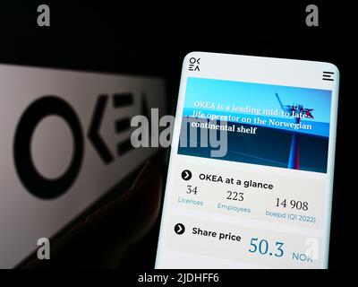 Person holding cellphone with website of Norwegian oil company OKEA ASA on screen in front of business logo. Focus on center of phone display. Stock Photo