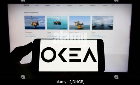 Person holding mobile phone with logo of Norwegian oil company OKEA ASA on screen in front of business web page. Focus on phone display. Stock Photo