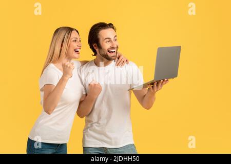Cheerful happy millennial caucasian couple in white t-shirts holding laptop, making victory sign with hands Stock Photo