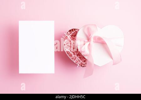 Top view of blank vertical paper card and white heart shape present box isolated on pastel pink background. Greeting concept. Stock Photo