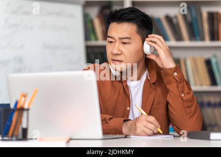 Asian Teacher Man Using Laptop Wearing Headphones In Classroom Indoor Stock Photo