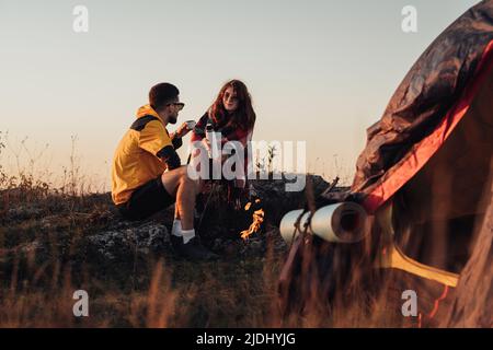 https://l450v.alamy.com/450v/2jdhyjg/two-young-travelers-man-and-woman-sitting-near-the-campfire-and-tent-outdoors-during-sunset-and-drinking-hot-tea-from-thermos-2jdhyjg.jpg