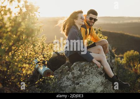 Happy Traveler Couple Man and Woman Sitting on the Edge of Rock During Sunset and Smiling, Wind Blowing Woman's Hair Stock Photo