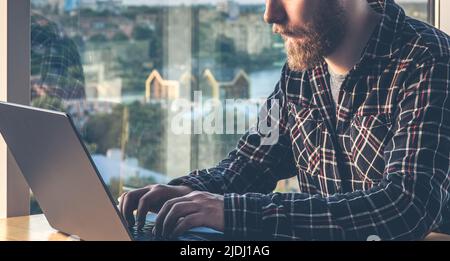 A man with a beard works at a computer, sitting in the office by the window. Stock Photo