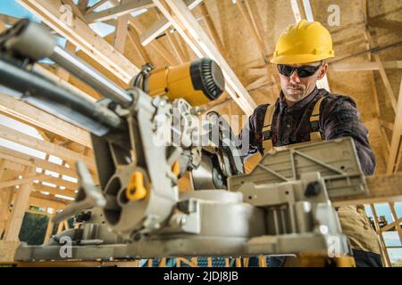Caucasian Worker Using a Table Saw While Working on the Construction of the Wooden Skeleton of a Residential Building. Construction Site Background. W Stock Photo