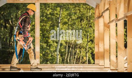 Construction Contractor Worker Wearing Hard Hat with Nail Gun in His Hand Walking Along Wooden Skeleton Frame Roof Beam. Construction Industry Theme.