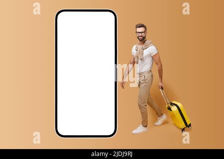 Mobile app for travelers. Handsome man with yellow luggage suitcase walking to huge smartphone with empty display, copy space, mockup Stock Photo