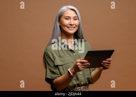 Modern elderly senior woman using digital tablet isolated on brown. Portrait of mature female office employee using online technology for doing business, computer app for accounting Stock Photo