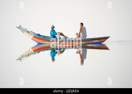 Two young men, one rowing and one on mobile phone in a brightly painted wooden boat perfectly reflected in mirror surface water Stock Photo