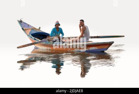 Two young men, one rowing and one on mobile phone in a brightly painted wooden boat perfectly reflected in mirror surface water Stock Photo
