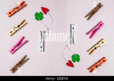 Colorful set of different wooden and plastic clothes pegs on a light pink glitter background. A red and a green lucky clover leaf are attached to each Stock Photo