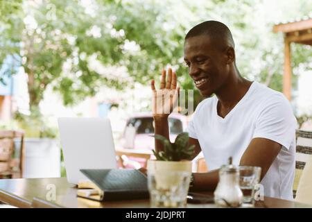 Smiling and glad multiracial man with laptop, raising arm, greet someone, looking at monitor. E-learning webinar meeting Stock Photo