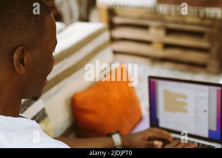 Rearview concentrated african afro american man with laptop sit on bench with legs in outdoor cafe. Editing contracts Stock Photo