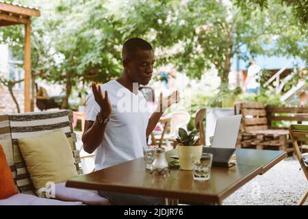 Surprised and confused african afro american man with laptop in cafe dont know what to do, raising hands in outdoor cafe Stock Photo