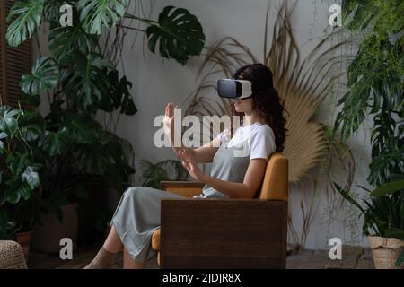 Young woman wearing VR helmet experiencing virtual reality while resting in greenhouse Stock Photo
