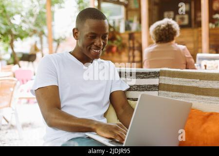 Concentrated multiracial man with laptop sitting on bench with legs in street coffee shop. Preparing to online webinar Stock Photo