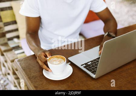 Unrecognizable cropped multicultural man using laptop, taking notes in workbook, drink coffee during internship study Stock Photo