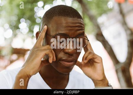 exhausted, stressed multiracial dark skin man in glasses keeping hands on head, suffering, screwing up eyes Stock Photo