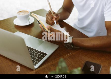 Unrecognizable cropped multicultural man using laptop, taking notes in workbook, drink coffee during internship study Stock Photo
