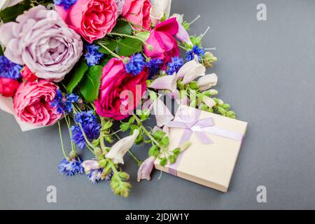 Close up of summer bouquet of roses foxgloves bachelor buttons flowers arranged on grey background with gift box. Surprise for holiday Stock Photo