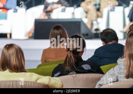 Back view of anonymous male and female participants in formal outfits sitting in armchairs and listening to crop speakers during business seminar in m Stock Photo