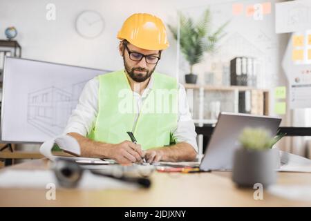 Architect man or male builder hold pen and ruler, writing and drawing architectural project, work on laptop computer on the desk at workplace. Stock Photo