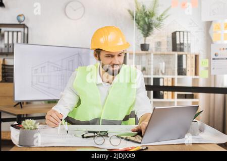 Competent architect male sitting on desk indoors with digital laptop using compass for project drawings. Positiv bearded young man wearing uniform green reflective vest, and construction hard hat. Stock Photo