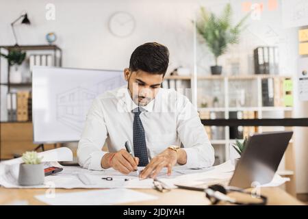 Bearded engineer indian man in yellow helmet and reflective vest sitting at table with lots of blueprints on the background of large tv screen with sketch. Business man working on construction project Stock Photo
