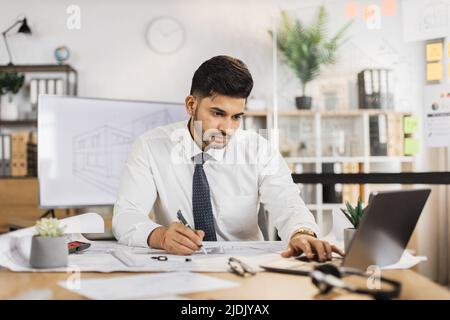 Bearded engineer indian man in yellow helmet and reflective vest sitting at table with lots of blueprints on the background of large tv screen with sketch. Business man working on construction project Stock Photo