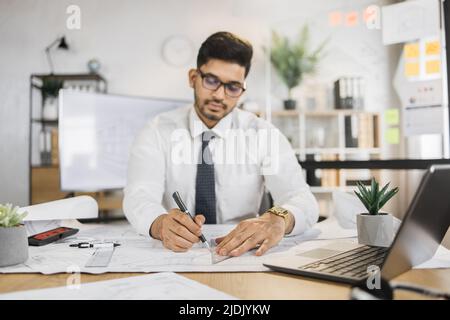 Portrait of indian man wearing formal wear and hard hat at work in modern office. Confident contractor sitting at the desk, holding pen and working on laptop with sketches of building object. Stock Photo