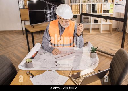 Competent architect senior male sitting on desk indoors with digital laptop using pen for project drawings. Positiv bearded mature man wearing orange reflective vest, and construction hard hat. Stock Photo
