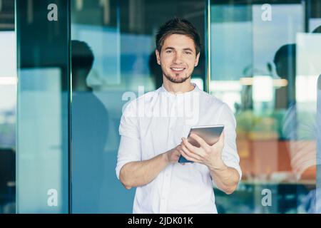 Portrait of a young handsome male student. Stands using a tablet, studying, reading. He looks at the camera, smiles Stock Photo