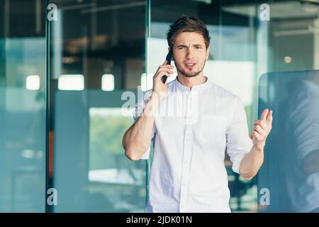 Worried young businessman talking on cell phone near office. Waving his arms, dissatisfied Stock Photo