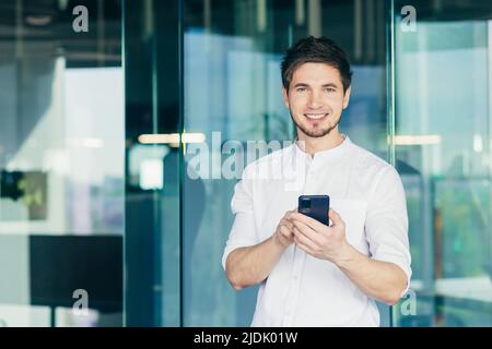 Young handsome male businessman standing in office, using cellphone. Gathers messages, calls. He stands, looks at the camera, smiles Stock Photo