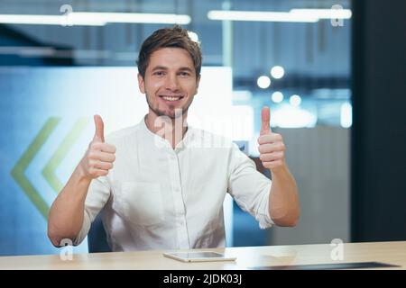 Portrait of a young successful handsome businessman, freelancer, manager, IT specialist. He sits at a table in the office, shows his hand super, looks Stock Photo