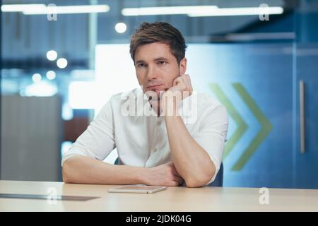 Portrait of a young male student. Serious, preparing for the exam, studying online, remotely. Sitting at the table with a laptop, tired, holding his h Stock Photo